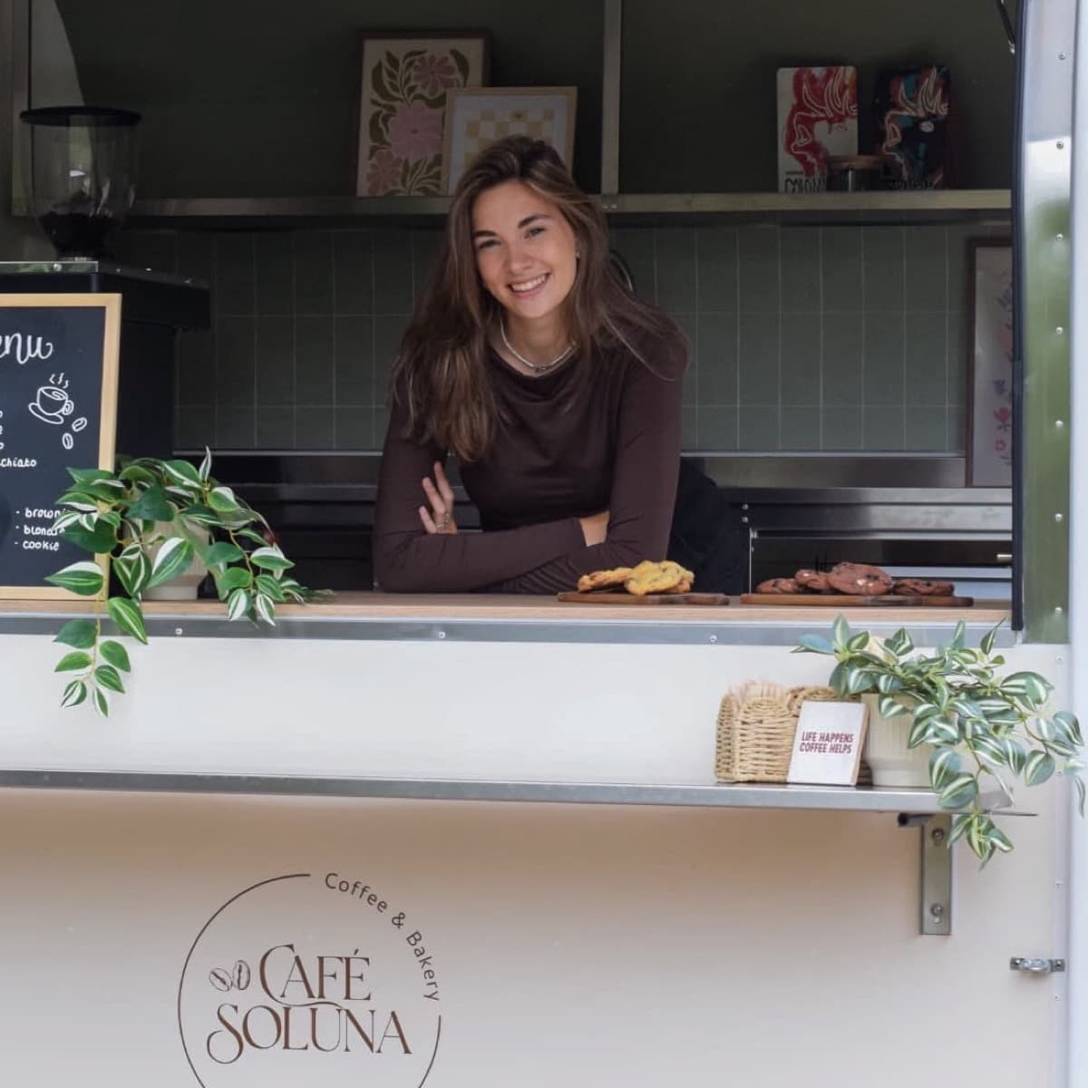 Melissa lacht vanuit het luik van de foodtruck, leunend op de toonbank, met het menubord naast haar en het logo op de wand eronder