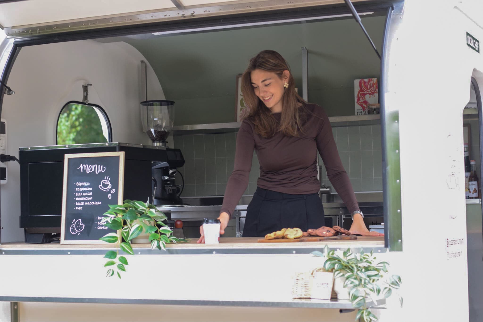 Melissa zet een beker koffie op de toonbank van de truck, met het menubord en vers gebak naast haar